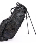 A stand golf bag with a dark camouflage pattern, featuring black triple legs and multiple zippered pockets, photographed on a solid white background.